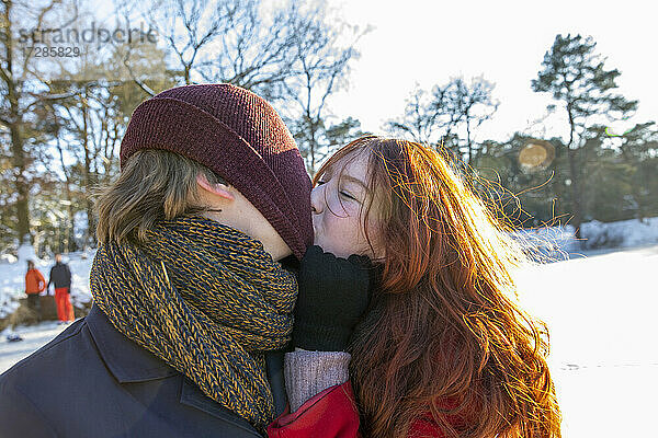 Rothaarige Frau zieht Strickmütze und küsst ihren Freund an einem sonnigen Tag