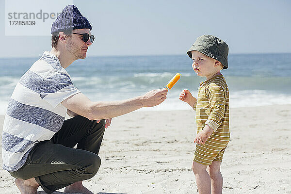 Mann mit Sonnenbrille füttert seinen Sohn am Strand mit Eiscreme