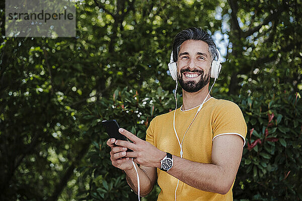 Lächelnder Mann mit Kopfhörern und Mobiltelefon im Park