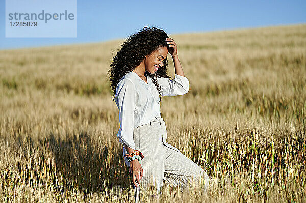 Lächelnde junge Frau mit Hand im Haar  die an einem sonnigen Tag durch ein Weizenfeld spaziert
