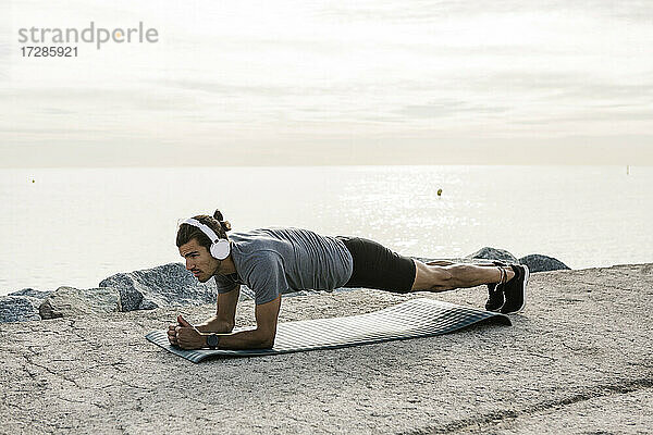 Männlicher Sportler mit Kopfhörern  der auf einer Trainingsmatte die Plank-Position übt