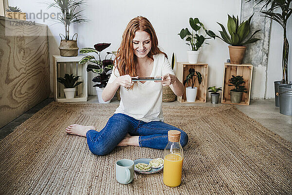 Lächelnde junge Frau fotografiert Essen  während sie zu Hause auf dem Boden sitzt