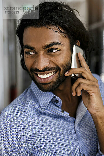Lächelnder junger männlicher Unternehmer  der im Büro mit dem Handy telefoniert und wegschaut