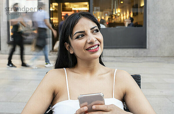 Lächelnde Frau träumt  während sie ihr Smartphone in einem Straßencafé hält