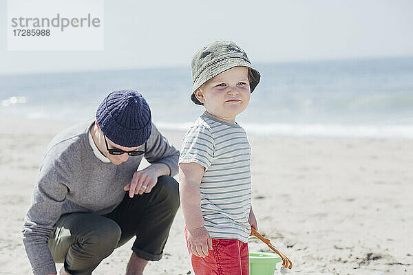 Niedlicher kleiner Junge  der wegschaut  während er neben seinem Vater am Strand steht