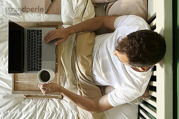 Mann sitzt mit Kaffeetasse und Laptop auf dem Tisch im Bett