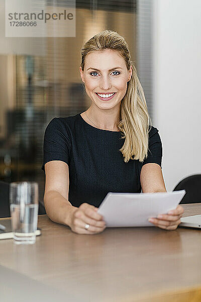 Schöne lächelnde Unternehmerin mit Dokument sitzt am Schreibtisch im Büro