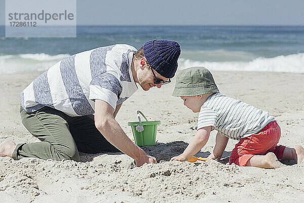 Lächelnder Mann gräbt mit seinem Sohn am Strand bei Sonnenschein im Sand