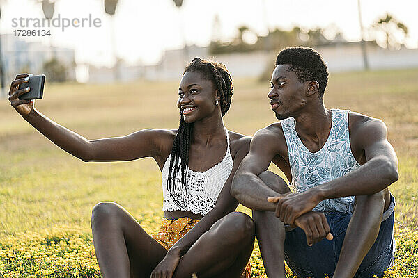 Paar nimmt Selfie durch Handy beim Sitzen auf Wiese während Sonnenuntergang