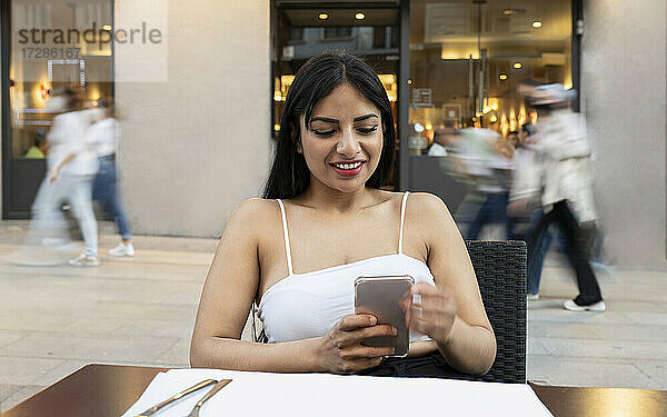 Lächelnde junge Frau  die in einem Straßencafé ein Mobiltelefon benutzt