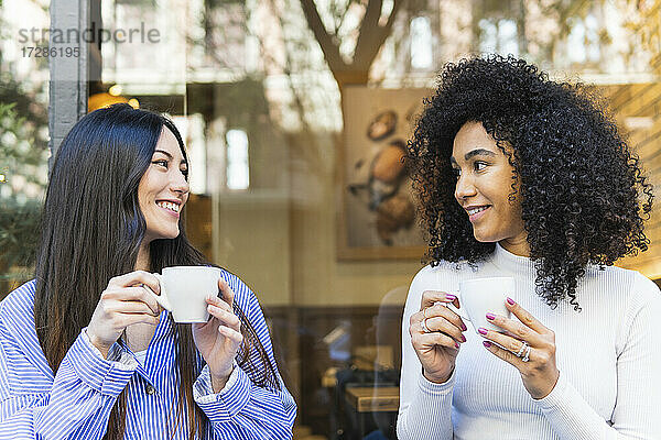 Lächelnde junge Frau sieht ihre Freundin an  während sie vor einer Bar Kaffee trinkt