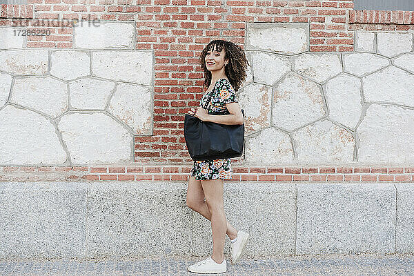 Lächelnde schöne Frau  die eine Handtasche trägt  während sie an einer Backsteinmauer steht
