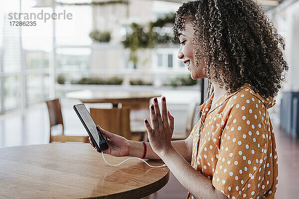 Lächelnde Frau grüßt während eines Videogesprächs über ein Mobiltelefon in einem Café