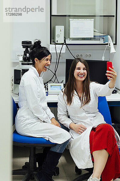 Fröhliche junge Medizinerin  die ein Selfie mit einem Kollegen im Labor über ihr Smartphone macht