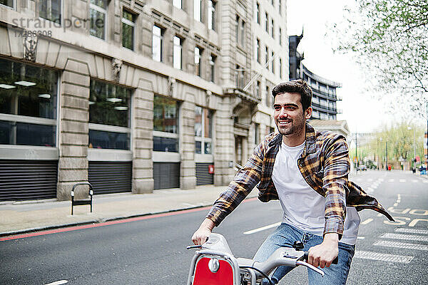 Lächelnder Mann mit kariertem Hemd  der auf einer Straße in der Stadt Rad fährt