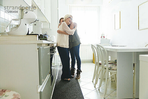 Ältere Frau und Enkelin umarmen sich  während sie in der Küche zu Hause stehen
