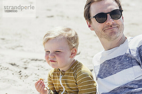 Lächelnder Mann mit Sonnenbrille sitzt mit seinem Sohn am Strand