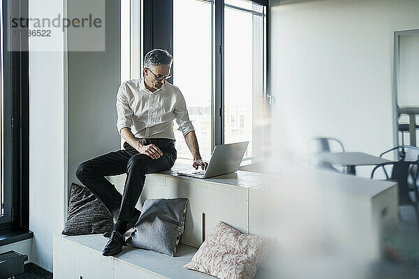 Männlicher Unternehmer  der einen Laptop benutzt  während er auf einem Möbelstück im Büro sitzt