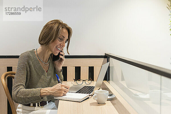 Frau mit Laptop  die in ein Buch schreibt  während sie in einem Café mit ihrem Handy telefoniert