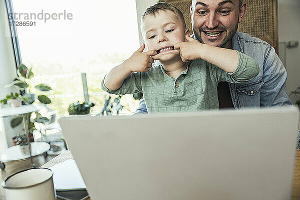 Junge neckt  während er mit seinem Vater sitzt und einen Videoanruf über den Laptop zu Hause tätigt