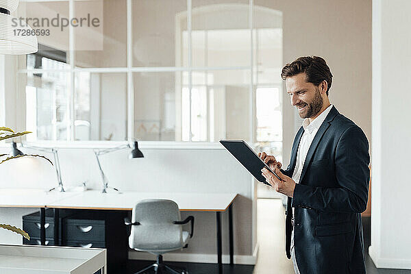 Älterer Geschäftsmann  der im Büro stehend ein digitales Tablet benutzt