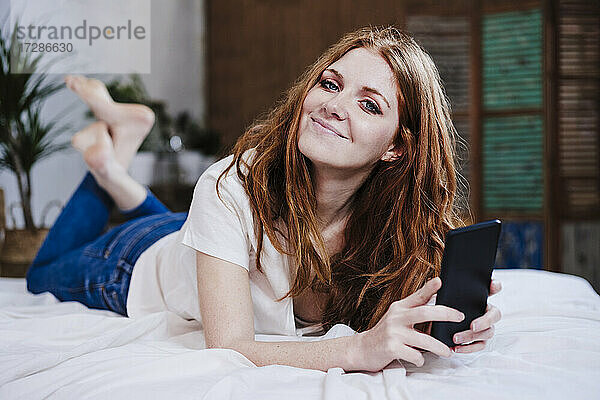 Schöne Frau lächelt und hält Handy auf dem Bett zu Hause