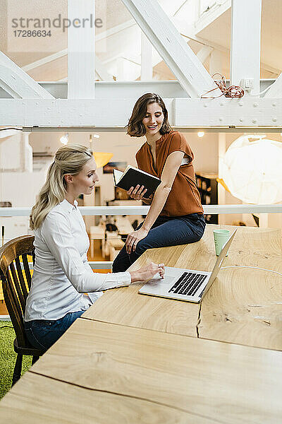 Lächelnde junge Geschäftsfrau  die auf ihren Laptop schaut  während sie mit einer Kollegin in einem kreativen Büro einen Plan bespricht