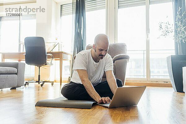 Junger Mann benutzt einen Laptop  während er zu Hause auf einer Übungsmatte sitzt