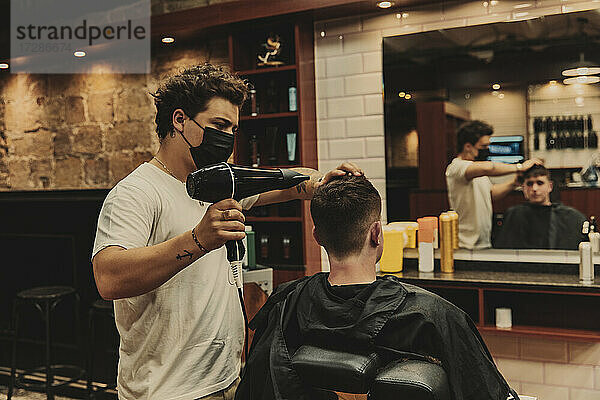 Männlicher Friseur mit Haartrockner für jungen Mann im Salon während COVID-19