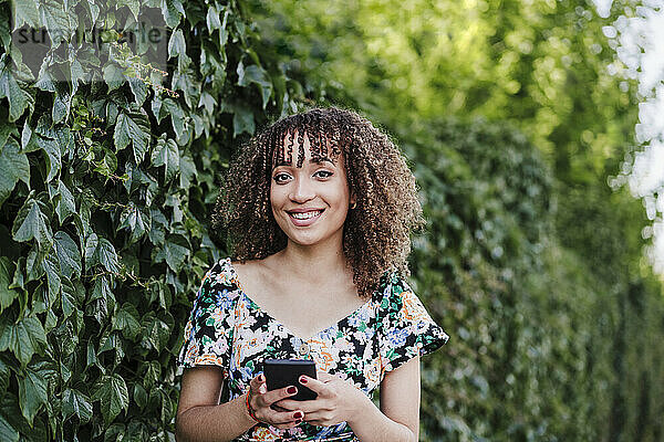 Lächelnde junge Frau  die ihr Smartphone in der Hand hält und neben einer Efeupflanze steht