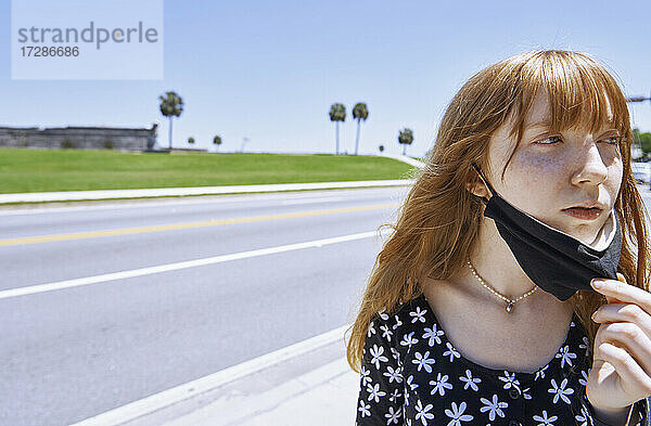 Rothaarige junge Frau mit Gesichtsschutzmaske bei COVID-19  die an der Straße steht und wegschaut