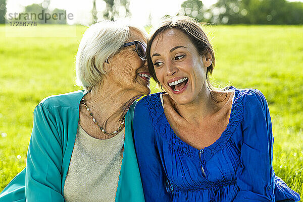 Ältere Frau flüstert mit einer Frau im mittleren Erwachsenenalter  während sie in einem öffentlichen Park sitzt