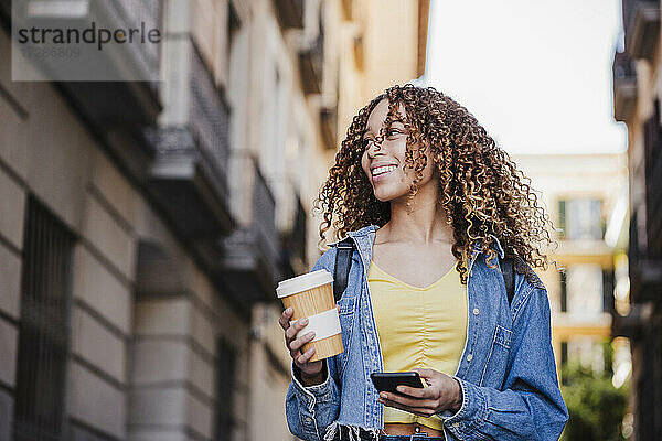 Schöne Frau schaut weg  während sie einen wiederverwendbaren Becher und ein Smartphone in der Stadt hält