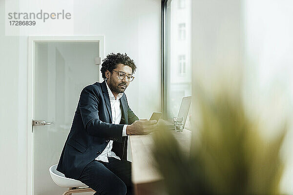 Männlicher Berufstätiger  der ein Smartphone benutzt  während er im Büro am Schreibtisch sitzt