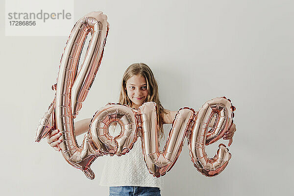 Lächelndes Mädchen zeigt Liebesballon  während sie vor einer Wand steht
