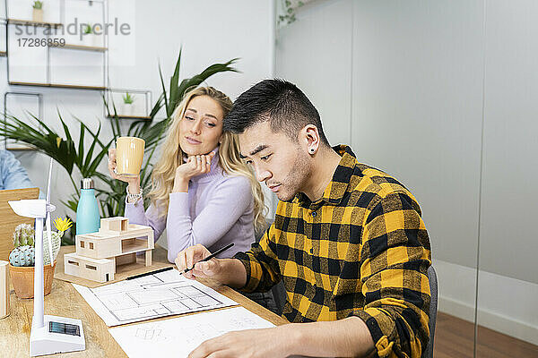 Ein männlicher Architekt fertigt einen Bauplan an  während eine Mitarbeiterin am Schreibtisch Kaffee trinkt.