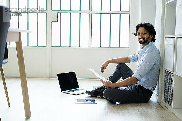 Männlicher Unternehmer mit digitalem Tablet und Laptop auf dem Boden sitzend im Büro