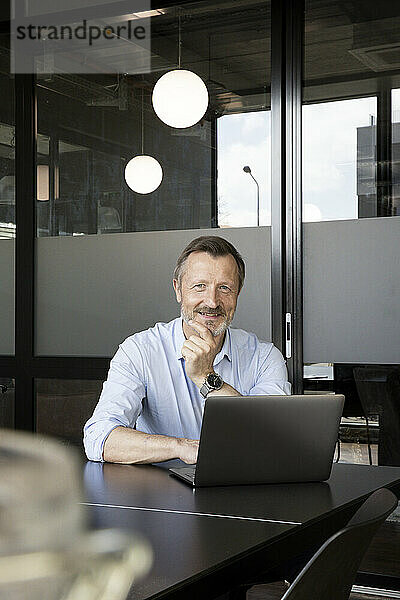 Männlicher Berufstätiger  der vor einem Laptop im Büro sitzt und lächelt