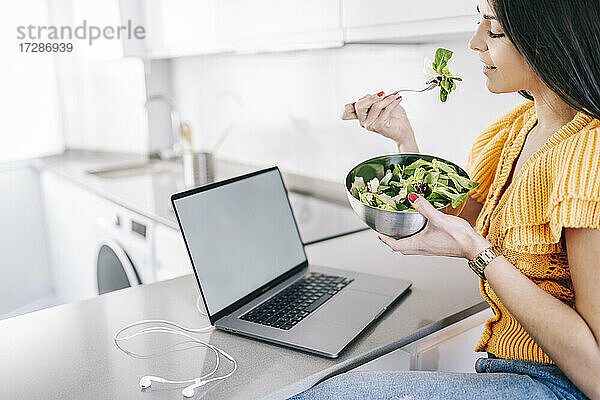 Frau isst Salat und sitzt am Laptop in der Küche