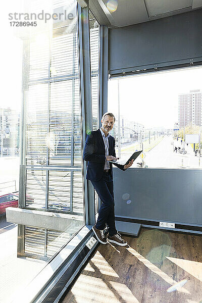 Männlicher Berufstätiger hält Laptop und lehnt sich an ein Glasfenster im Büro