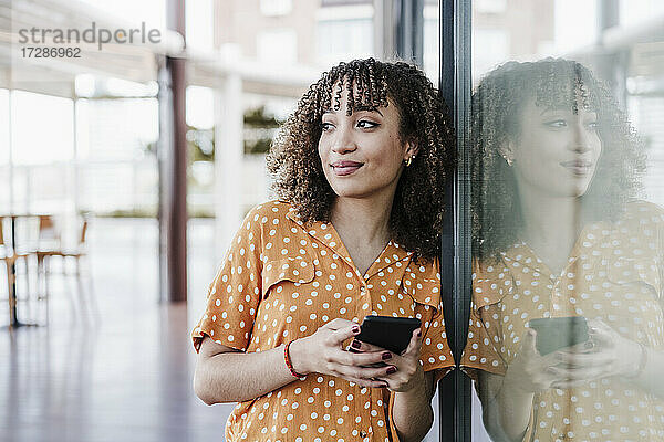 Schöne junge Frau mit Mobiltelefon  die sich an ein Glasfenster lehnt