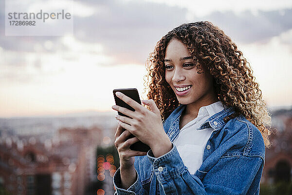 Lächelnde Frau mit Smartphone vor dem Himmel bei Sonnenuntergang