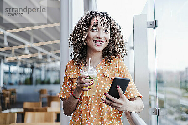 Lächelnde junge Frau mit Mobiltelefon  die wegschaut  während sie auf dem Balkon eines Cafés einen Eistee trinkt