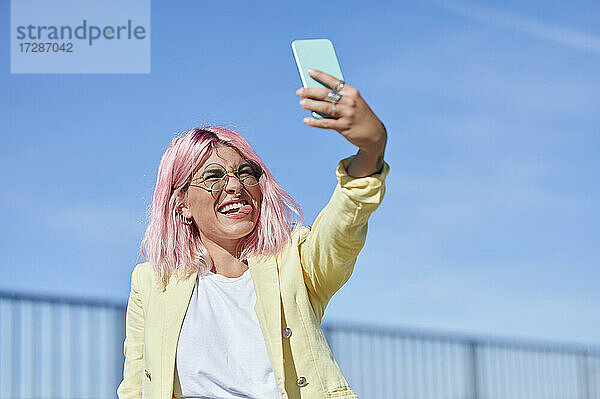Junge Frau streckt die Zunge heraus  während sie ein Selfie mit ihrem Smartphone an einem sonnigen Tag macht