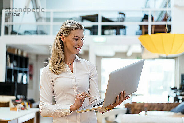 Lächelnde blonde Frau  die einen Laptop benutzt  während sie in einem kreativen Büro steht