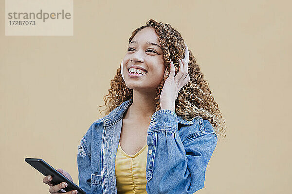 Lächelnde Frau hört Musik mit Kopfhörern an einer beigen Wand