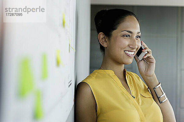 Lächelnde Geschäftsfrau  die wegschaut  während sie mit dem Handy im Büro telefoniert