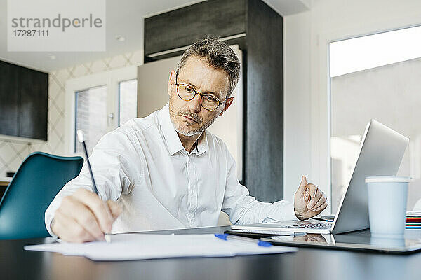 Älterer Geschäftsmann schreibt an einem Dokument  während er vor einem Laptop im Büro zu Hause sitzt