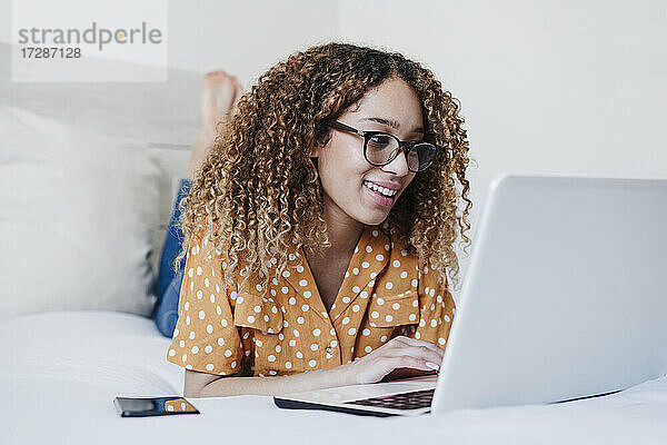 Junge Frau mit Brille benutzt einen Laptop  während sie zu Hause auf dem Bett liegt