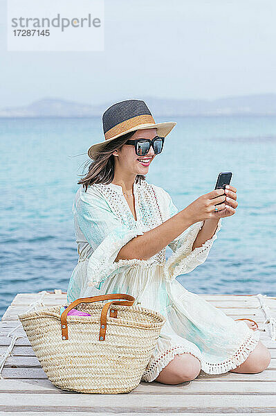 Lächelnde reife Frau mit Hut  die ein Selfie mit ihrem Smartphone macht  während sie auf dem Bootssteg sitzt